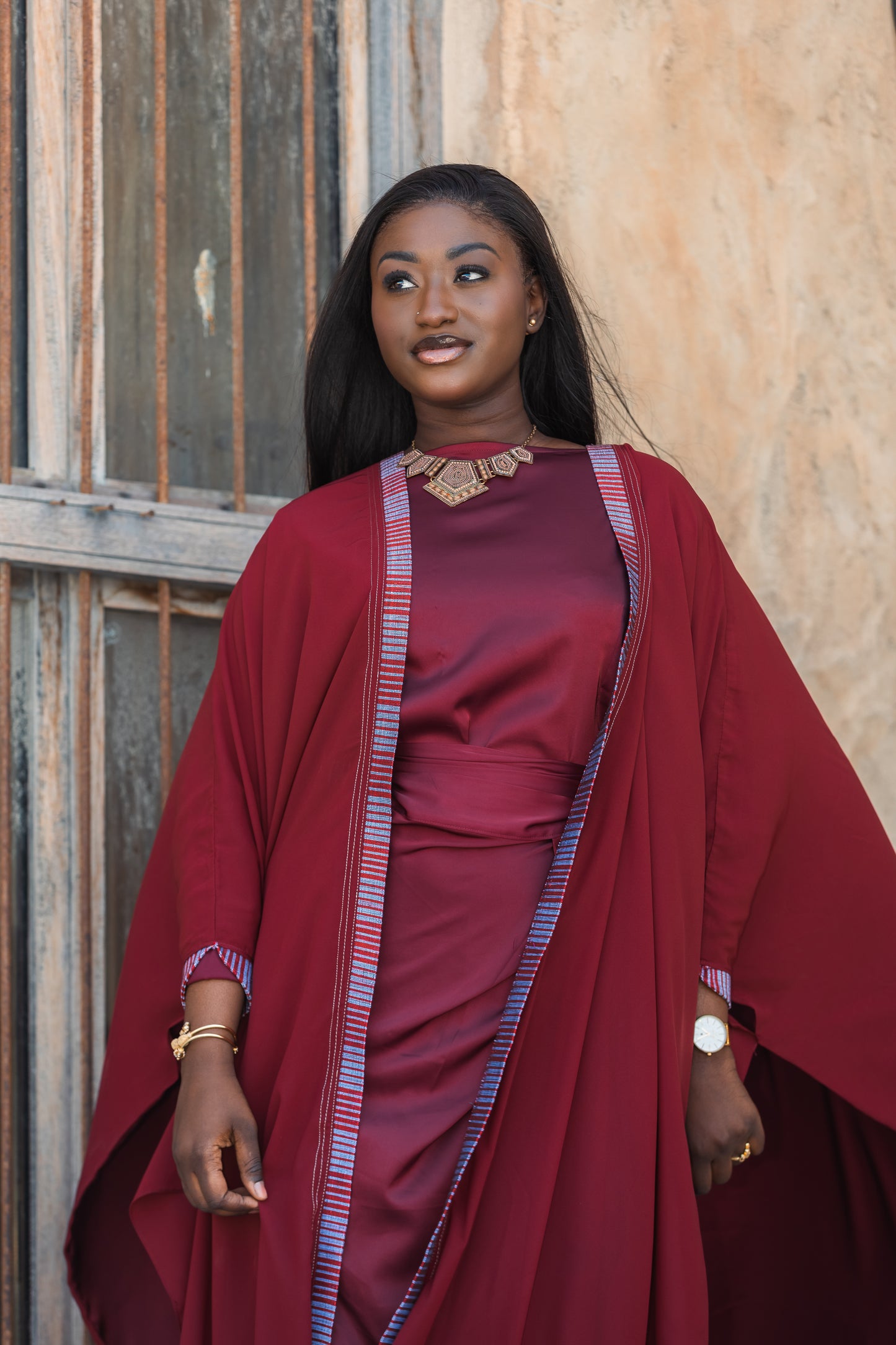 Woman wearing a red luxury abaya with a patterned shawl against a textured wall.