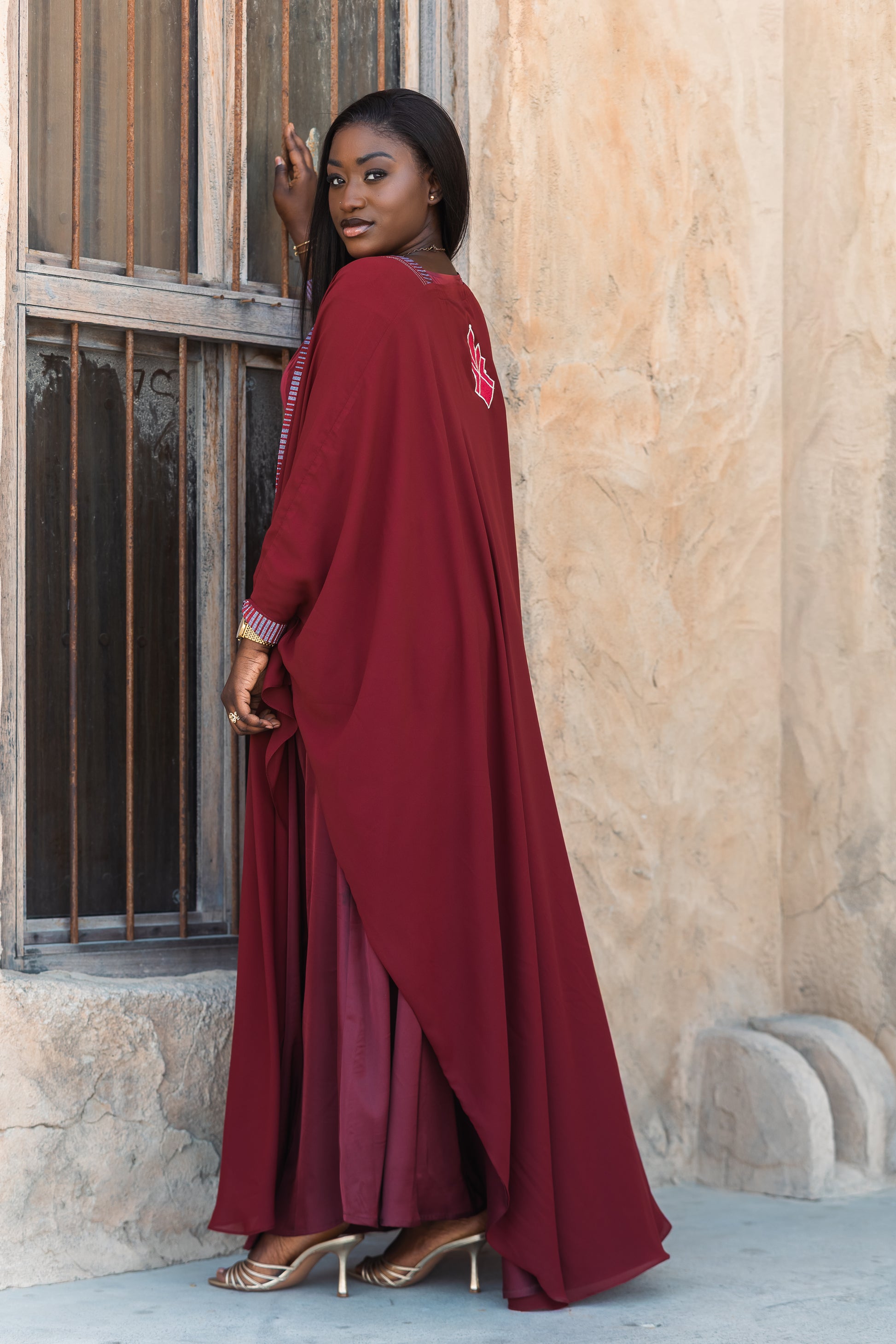 Woman wearing a maroon Eid abaya with artisan silk embroidery standing against a textured wall.