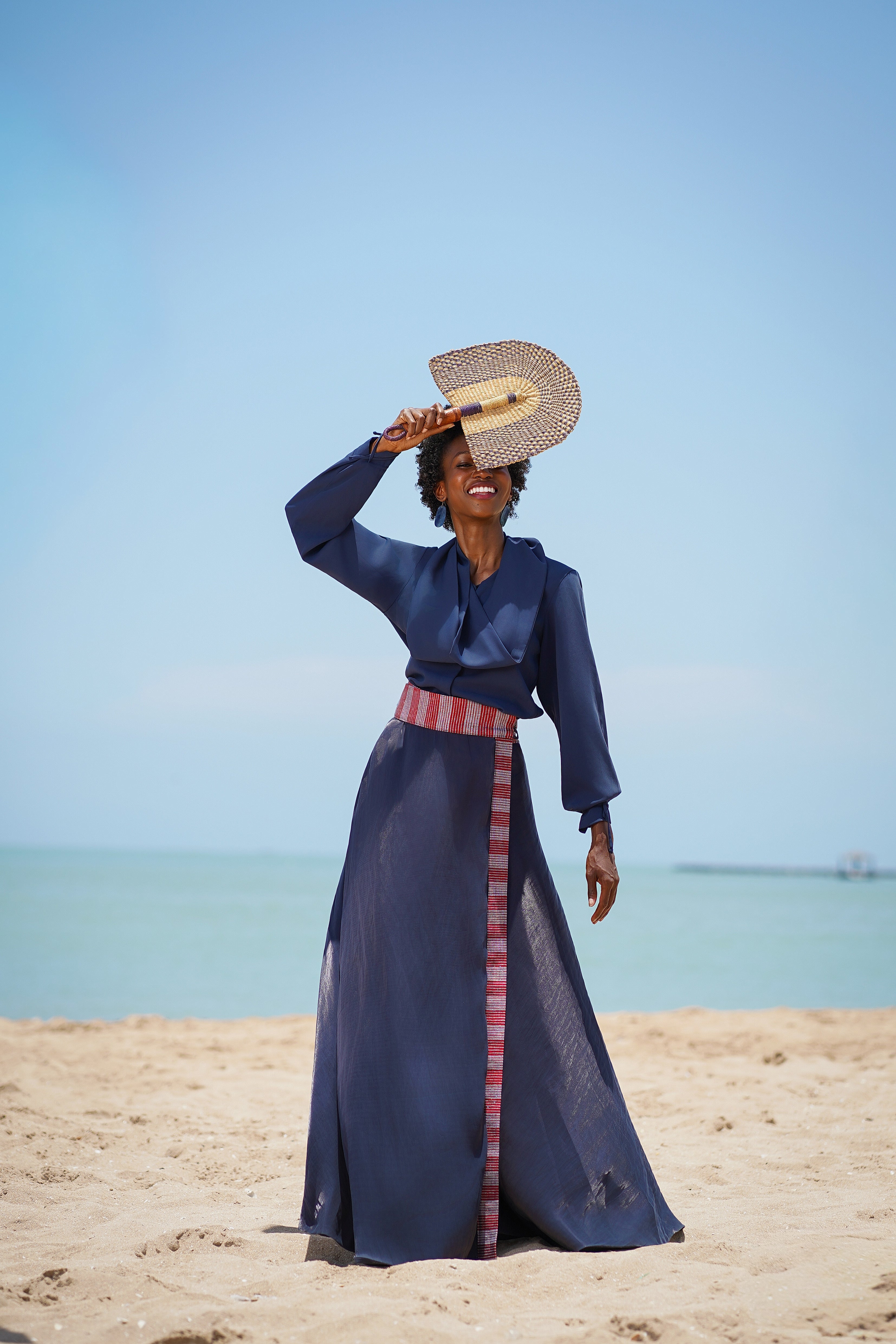 Woman in a blue dress with a striped belt standing on a beach with clear blue sky.